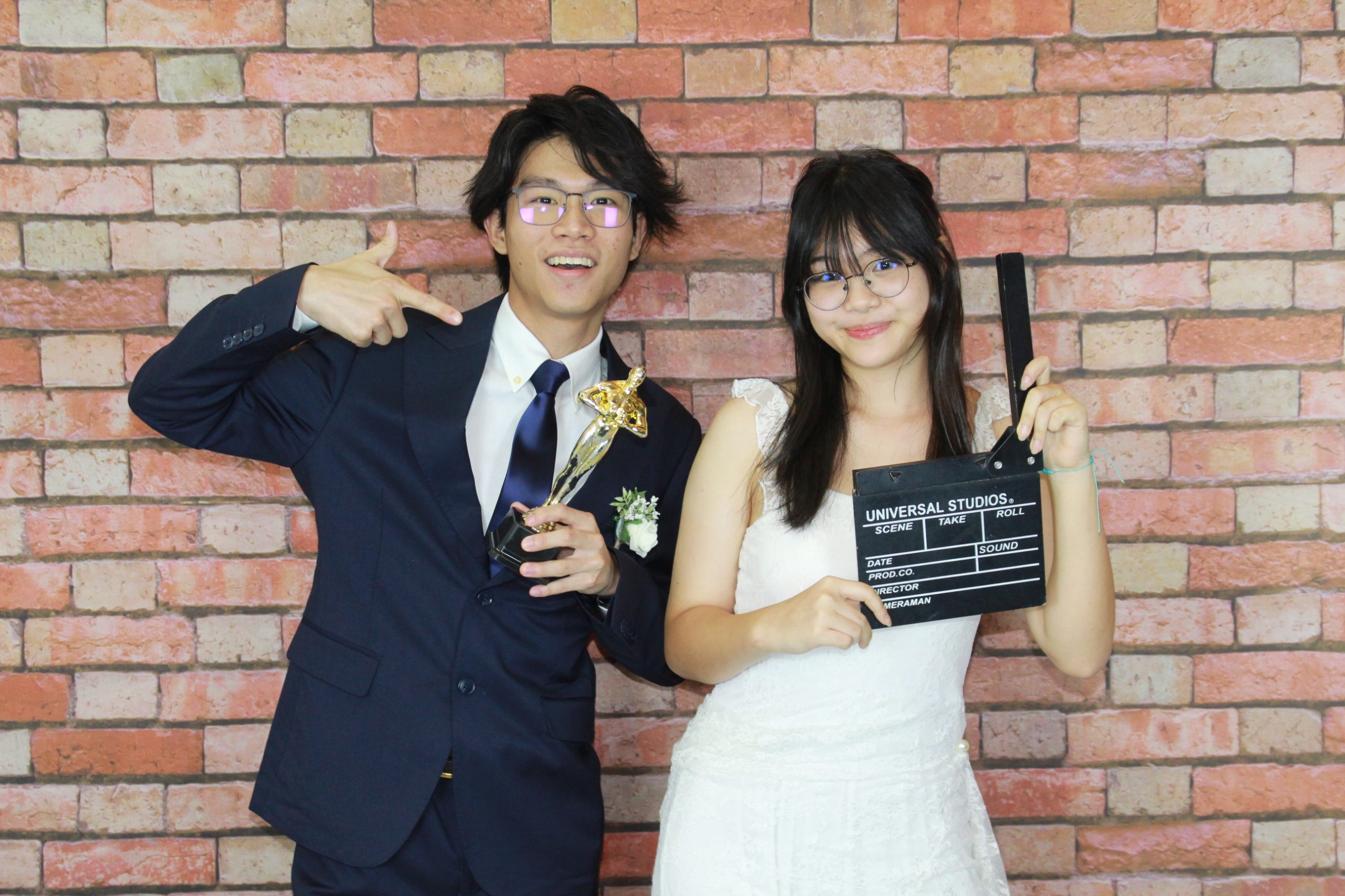 Wedding couple smiling with movie-themed props against a brick photo booth backdrop